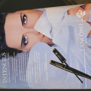 May include: A close-up of a woman's face with a white shirt draped over her. The woman is wearing mascara and has long black hair. The image also features two black mascara wands with gold accents and the text "Intencils Lash by Lash Full Intensity Mascara" and "Lanc&ocirc;me Paris".