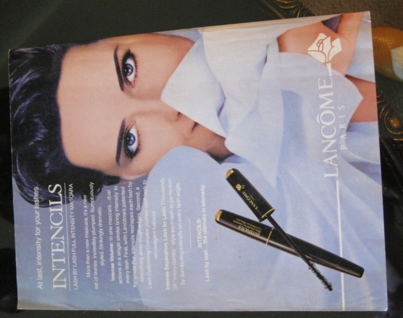 May include: A close-up of a woman's face with a white shirt draped over her. The woman is wearing mascara and has long black hair. The image also features two black mascara wands with gold accents and the text "Intencils Lash by Lash Full Intensity Mascara" and "Lanc&ocirc;me Paris".