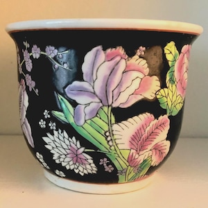 May include: A black ceramic plant pot with a white rim and floral design. The pot features large pink and purple flowers with green leaves. The flowers are painted on a black background.