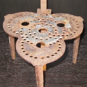 May include: A three-legged, clover-shaped metal trivet featuring a heavily rusted, weathered surface. The top plate is perforated with numerous small circular holes arranged in a repeating pattern, with three larger circular openings centered.