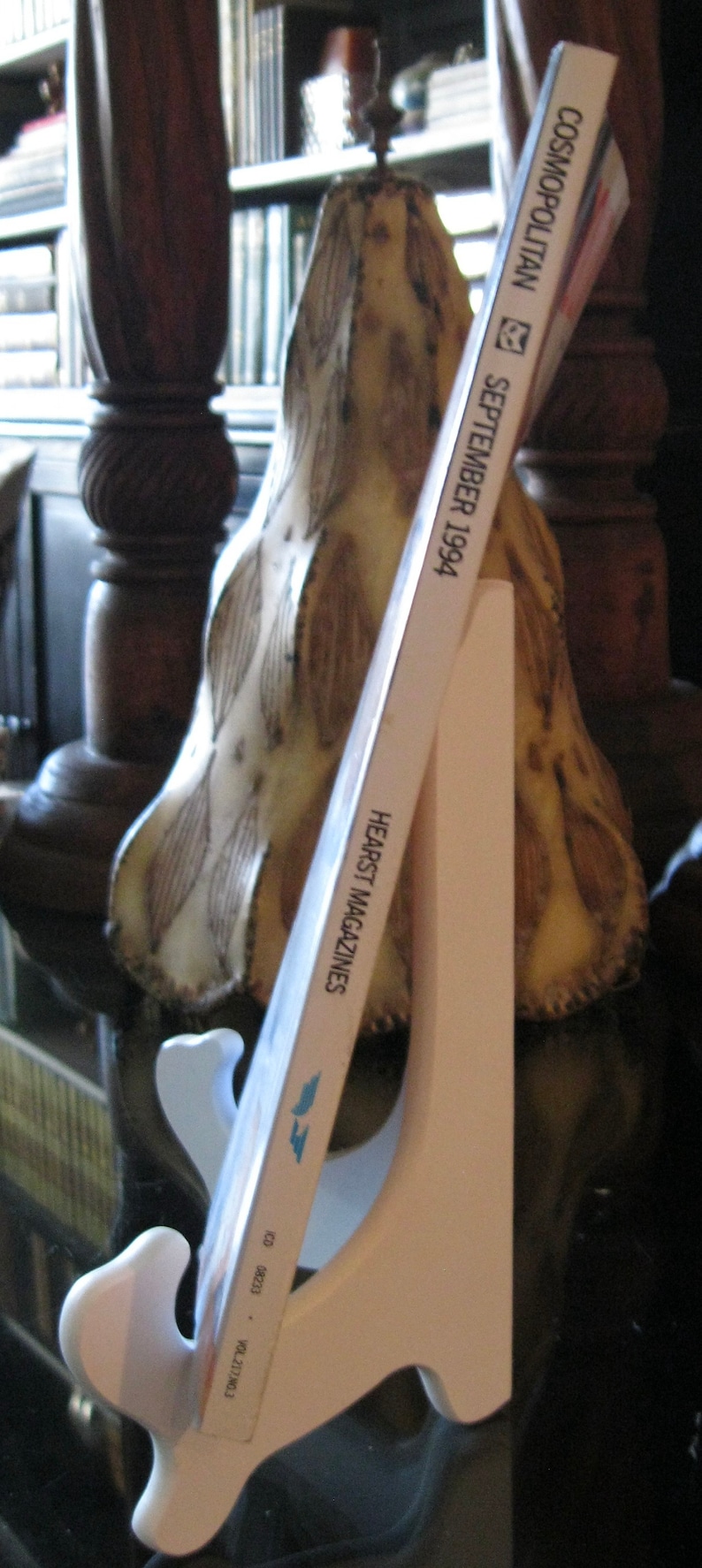 May include: A white magazine stand with a magazine titled "Cosmopolitan" September 1994, Hearst Magazines. The magazine is leaning against a decorative lamp with a white and brown patterned design.