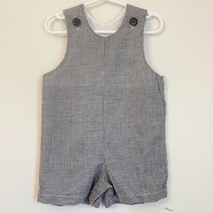 Può includere: Pantaloncini a salopette in vichy bianco e nero per un bambino o un bambino piccolo. I pantaloncini hanno uno sfondo bianco con un motivo a vichy nero e due bottoni neri sulle bretelle.