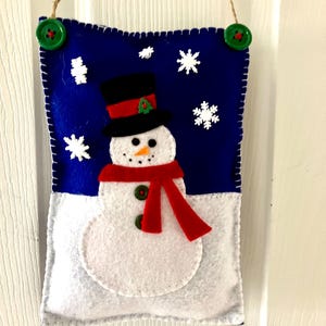 May include: A felt snowman ornament with a red scarf and black top hat hanging on a white door. The snowman is on a blue background with white snowflakes.