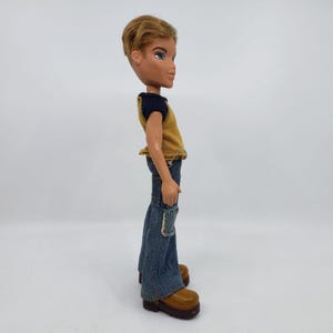 May include: A Bratz doll of a young man with short, styled blonde hair and blue eyes. He wears a yellow and navy blue top, blue denim jeans with patches, and brown platform boots. The doll is posed in profile.
