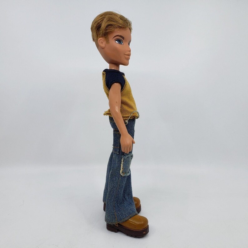 May include: A Bratz doll of a young man with short, styled blonde hair and blue eyes. He wears a yellow and navy blue top, blue denim jeans with patches, and brown platform boots. The doll is posed in profile.