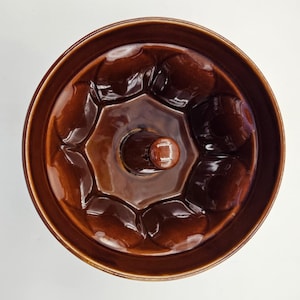 May include: A brown ceramic bundt pan with a central tube and a series of rounded indentations around the perimeter. The pan has a glossy finish and is designed for baking cakes. The pan is circular and sits on a white surface.