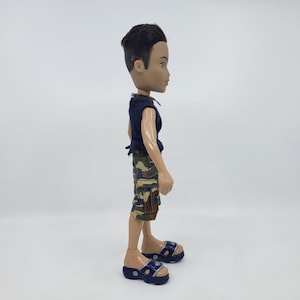 May include: A doll wearing a navy blue sleeveless shirt, camouflage shorts, and navy blue sandals. The doll has short black hair and is standing on a white surface. The doll is a Bratz doll.
