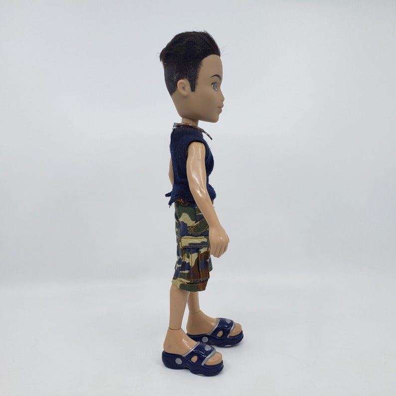 May include: A doll wearing a navy blue sleeveless shirt, camouflage shorts, and navy blue sandals. The doll has short black hair and is standing on a white surface. The doll is a Bratz doll.