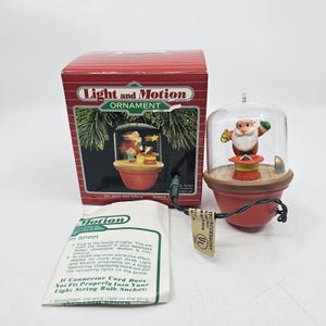 May include: A red Light and Motion Christmas ornament featuring Santa Claus inside a clear dome. The ornament is on a wooden base and has a red pot-shaped bottom. The box reads "Light and Motion Ornament".
