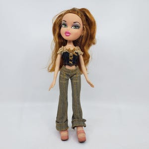 May include: A Bratz doll with long auburn hair and green eyes. She wears a black and tan corset top, plaid pants, and pink platform shoes. The doll is standing against a white background.