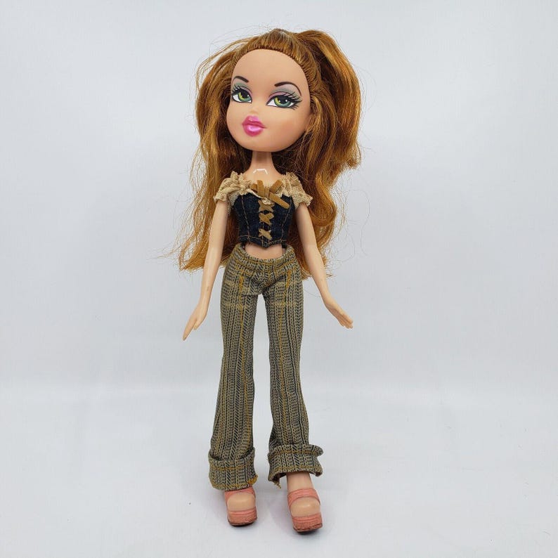 May include: A Bratz doll with long auburn hair and green eyes. She wears a black and tan corset top, plaid pants, and pink platform shoes. The doll is standing against a white background.