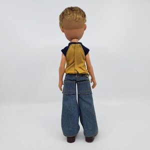 May include: A vintage doll with short, light brown hair and a tan complexion. The doll is wearing a yellow and navy blue top and blue denim jeans. The doll's brown shoes are visible.