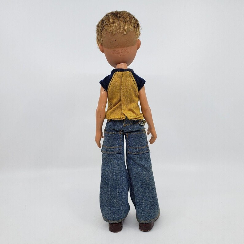 May include: A vintage doll with short, light brown hair and a tan complexion. The doll is wearing a yellow and navy blue top and blue denim jeans. The doll's brown shoes are visible.