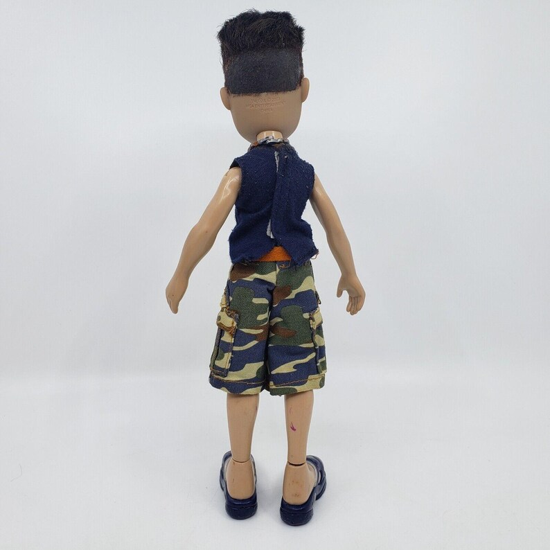 May include: A doll wearing a navy blue vest, orange undershirt, and camouflage cargo shorts. The doll has dark hair and is wearing navy blue sandals. The doll is standing against a white background.