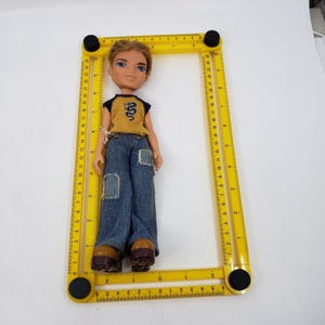May include: A Bratz doll with blonde hair, blue eyes, and a mustard yellow and navy blue top with a dragon design. The doll is wearing blue jeans with patches and brown shoes. A yellow measuring tool surrounds the doll.