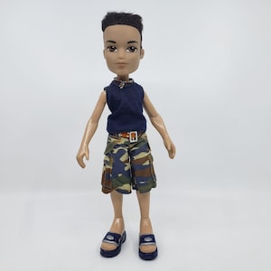 May include: A Bratz doll wearing a navy blue sleeveless shirt, camouflage shorts, and blue sandals. The doll has dark hair and is wearing a brown belt. The doll is standing against a white background.