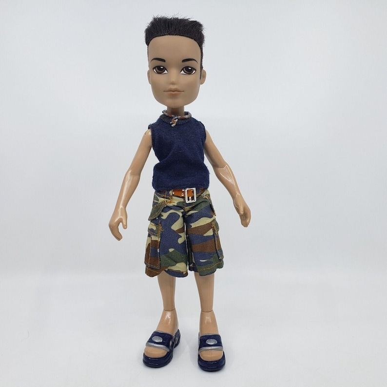 May include: A Bratz doll wearing a navy blue sleeveless shirt, camouflage shorts, and blue sandals. The doll has dark hair and is wearing a brown belt. The doll is standing against a white background.