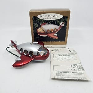 May include: A red and silver airplane ornament with a clear cockpit, part of the Hallmark Keepsake collection. The ornament is in its original box, with a warranty card. The airplane has a green hanging string.
