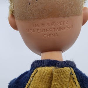 May include: A doll's head and shoulders are visible, with short, light brown hair and a tan complexion. The back of the head has the text "TM, &reg; & &copy; 2002 MGA ENTERTAINMENT CHINA". The doll is wearing a blue and yellow shirt.