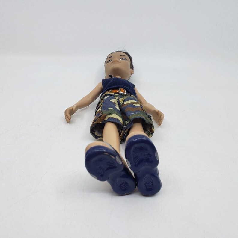 May include: A doll wearing a navy blue tank top and camouflage shorts with a brown belt. The doll has dark blue shoes. The doll is lying down on a white surface.