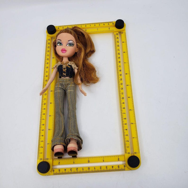 May include: A Bratz doll with auburn hair, wearing a black top, patterned pants, and pink shoes. The doll is positioned within a yellow measuring frame, with measurements in inches. The doll is on a white surface.