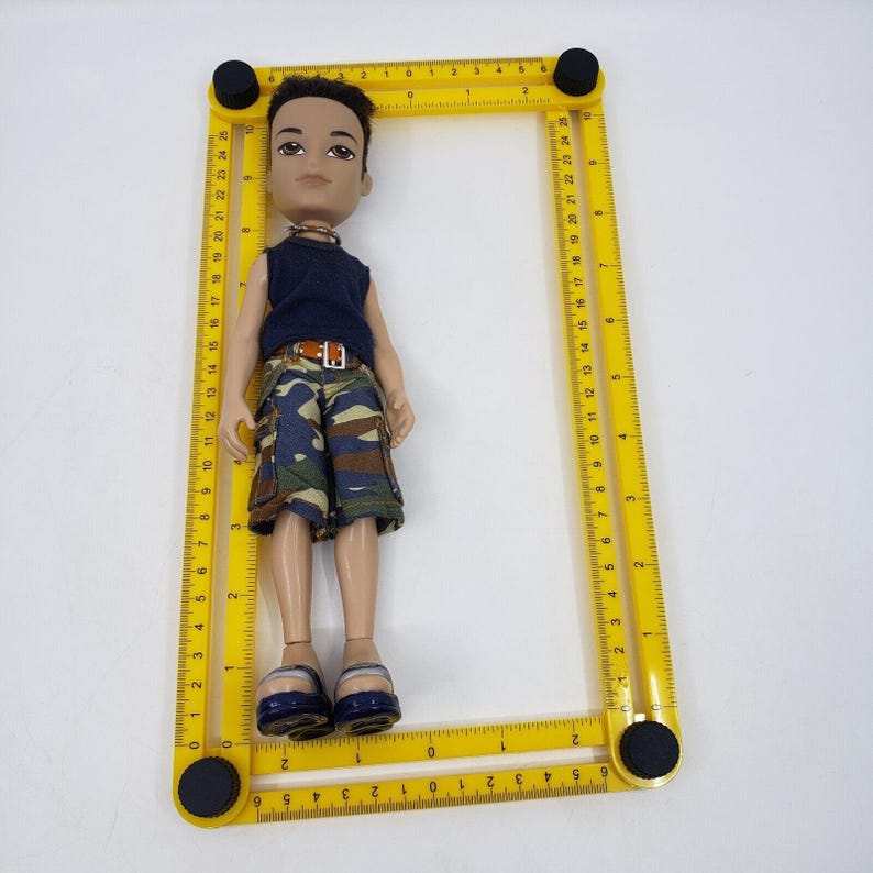 May include: A Bratz doll wearing a navy blue sleeveless shirt, camouflage shorts, and blue sandals. The doll is positioned inside a yellow adjustable ruler with black knobs. The ruler is marked with inch measurements.