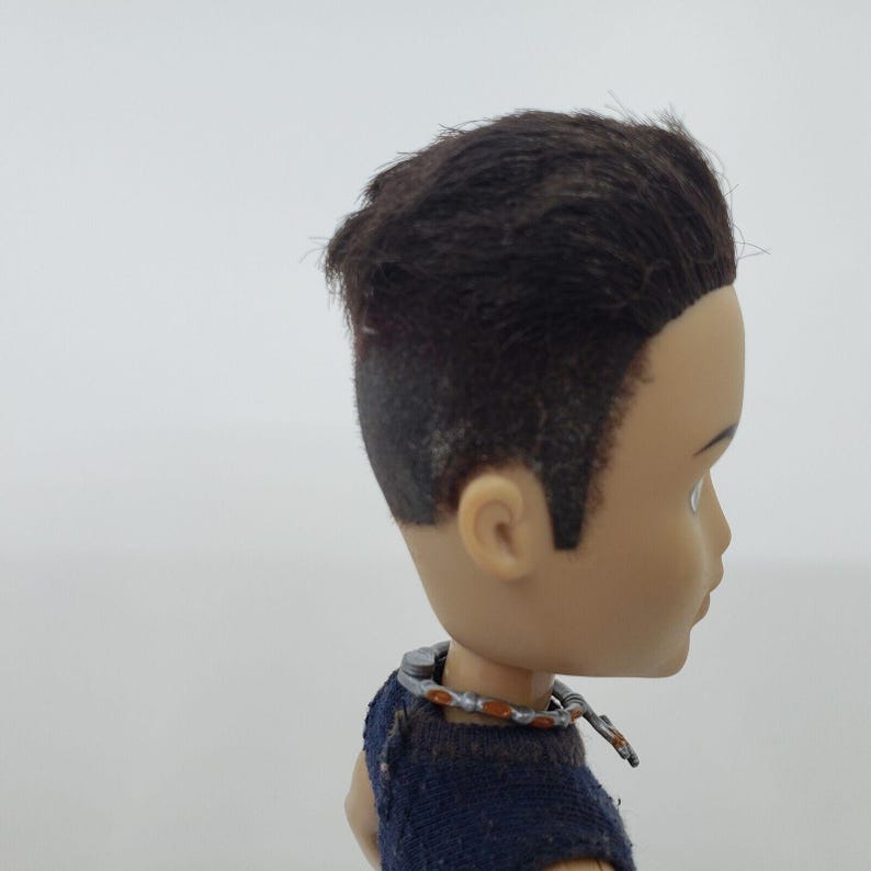 May include: A doll's head in profile, featuring dark brown hair with a faded undercut. The doll has a fair complexion and is wearing a blue top and a silver and orange necklace. The background is plain white.