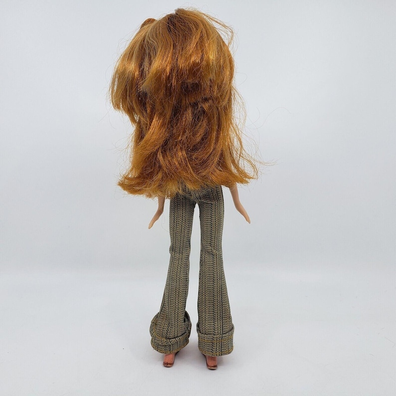 May include: A doll with long, red hair and wearing patterned, flared pants. The doll's back is facing the viewer, showcasing the hairstyle and the design of the pants. The doll's feet are visible, with small, pink shoes.