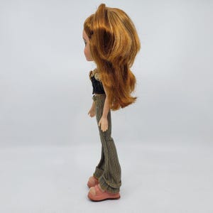 May include: A Bratz doll with auburn hair, wearing a black top, brown patterned pants, and pink sandals. The doll is posed in profile against a white background. The doll's hair is styled in a half-up, half-down fashion.