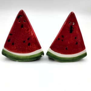 May include: A set of two ceramic watermelon salt and pepper shakers. The shakers are shaped like a slice of watermelon with red flesh, black seeds, and a green rind.