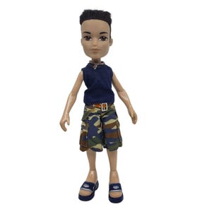 May include: A Bratz doll wearing a navy blue sleeveless shirt, camouflage shorts with a brown belt, and blue sandals. The doll has dark hair and is standing against a white background. The doll is a collectible toy.