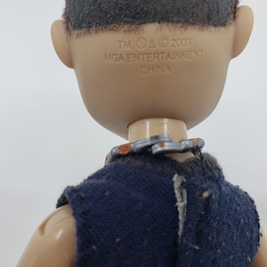 May include: Close-up of a doll's head and upper body. The doll has tan skin, black hair, and is wearing a dark blue top. The text "TM, &reg; & &copy; 2003 MGA ENTERTAINMENT CHINA" is printed on the back of the doll's head.