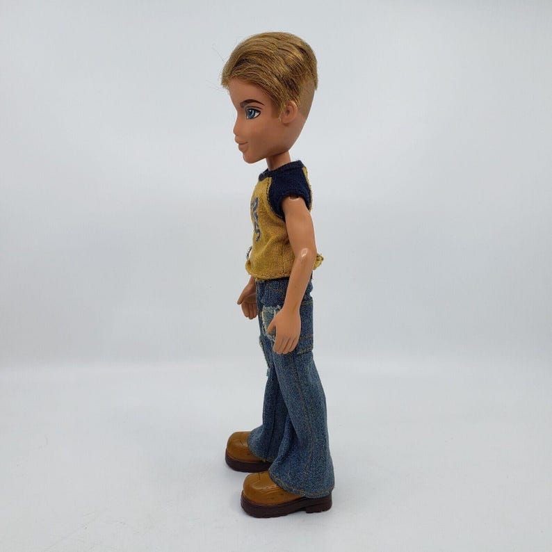 May include: A Bratz doll in a side profile view. The doll has short, light brown hair and blue eyes. It wears a yellow and navy blue shirt, ripped blue jeans, and brown boots. The doll is standing against a white background.
