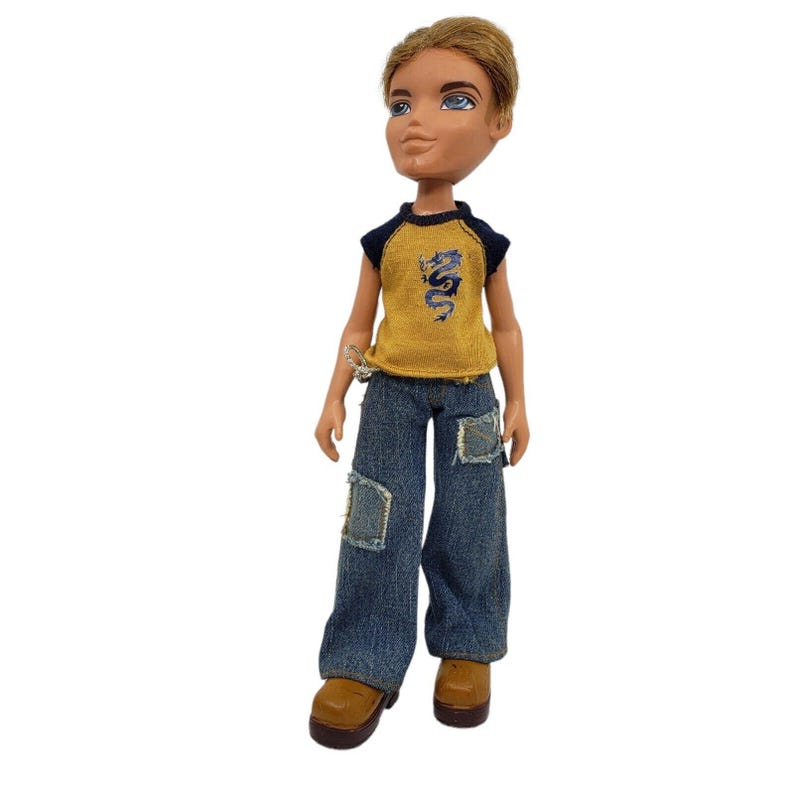 May include: A Bratz doll with short, light brown hair and blue eyes. The doll wears a yellow and navy blue sleeveless shirt with a dragon graphic, blue jeans with patches, and brown boots. The doll is standing.