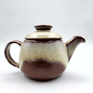 May include: A ceramic teapot with a lid. The teapot is a rounded shape with a handle and spout. The colors are brown, tan, and cream, with a gradient effect. The teapot is likely for brewing tea.