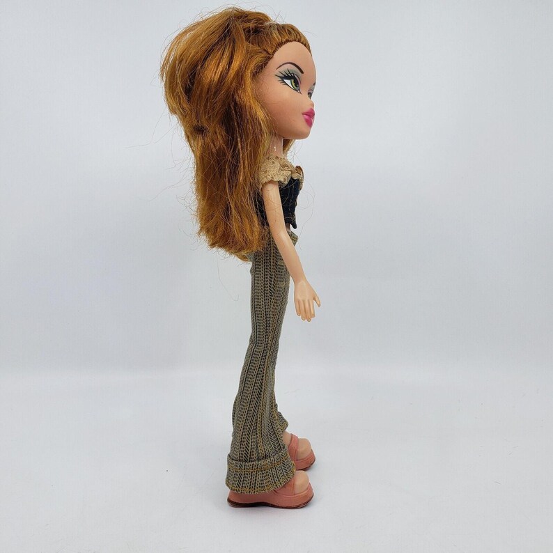 May include: A Bratz doll with auburn hair styled in a high ponytail. The doll wears a black top with a tan collar, brown patterned pants, and pink platform shoes. The doll is posed in a side profile against a white background.