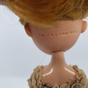 May include: Close-up of a doll's head and shoulders. The doll has blonde hair and a tan complexion. The back of the doll's head has the text "TM, R, and C 2001 MGA". The doll is wearing a beige lace top.