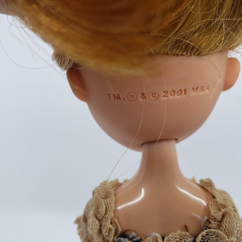 May include: Close-up of a doll's head and shoulders. The doll has blonde hair and a tan complexion. The back of the doll's head has the text "TM, R, and C 2001 MGA". The doll is wearing a beige lace top.