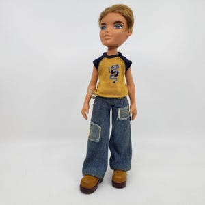 May include: A Bratz doll with short, light brown hair and blue eyes. The doll wears a mustard yellow t-shirt with a blue dragon graphic, blue denim jeans with patches, and brown boots. The doll is standing.
