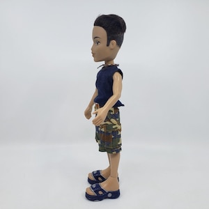 May include: A fashion doll with dark hair and tan skin, wearing a navy blue sleeveless top and camouflage print shorts. The doll is also wearing blue sandals. The doll is standing in profile against a white background.