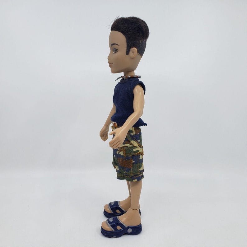 May include: A fashion doll with dark hair and tan skin, wearing a navy blue sleeveless top and camouflage print shorts. The doll is also wearing blue sandals. The doll is standing in profile against a white background.