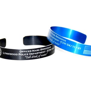 May include: Two engraved metal bracelets, one black and one blue. The black bracelet has a police badge and the text "OFFICER MARK BRINKMAN LYNNWOOD POLICE DEPARTMENT - EOW 04 "Tall drink of water"". The blue bracelet has the text "OFFICER MARK BRINKMAN DEPARTMENT - EOW 04/11/21 "Tall drink of water" and an American flag.