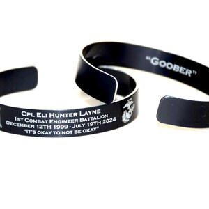 May include: Two black metal bracelets. One bracelet has an inscription that reads "CPL ELI HUNTER LAYNE 1ST COMBAT ENGINEER BATTALION DECEMBER 12TH 1999-JULY 19TH 2024 "IT'S OKAY TO NOT BE OKAY" ". The other bracelet has the inscription "GOOBER".