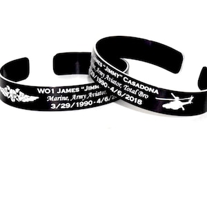 May include: Two black metal cuff bracelets with white text and graphics. One bracelet reads "WO1 JAMES 'JIMMY' Marine, Army Aviator" with dates and an emblem. The other reads "JAMES 'JIMMY' CASADONA, Army Aviator, Total Bro" with dates and a helicopter graphic.