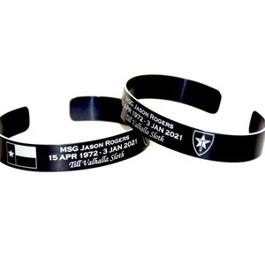 May include: Two black metal bracelets with engraved text. The top bracelet has a star and a sword design. The bottom bracelet has a Texas flag and the text "MSG JASON ROGERS 15 APR 1972-3 JAN 2021 Till Valhalla Sloth".