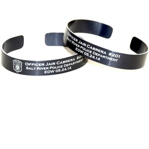 May include: Two black metal bracelets with white engraved text. The text reads "OFFICER JAIR CABRERA #201 SALT RIVER POLICE DEPARTMENT EOW 05.24.14" and "OFFICER JAIR CABRERA #2 SALT RIVER POLICE DEPARTMENT EOW 05.24.14".