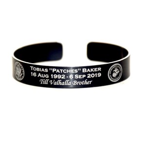May include: Black metal bracelet with a white inscription that reads "Tobias "Patches" Baker 16 Aug 1992 - 6 Sep 2019 Till Valhalla Brother". The bracelet features a US Army and US Marine Corps emblem.