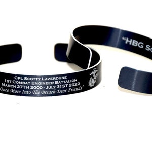 May include: Two black metal bracelets. One bracelet has an inscription that reads "CPL SCOTTY LAVERDURE 1ST COMBAT ENGINEER BATTALION MARCH 27TH 2000-JULY 31ST 2022 'Once More Into The Breach Dear Friends'" and a Marine Corps emblem. The other bracelet has the inscription "HBG SCOOT".