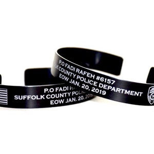 May include: Two black metal cuff bracelets. One bracelet features the American flag and the text "P.O FADI RAFEH #6157 SUFFOLK COUNTY POLICE DEPARTMENT EOW JAN. 20, 2019". The other bracelet has the same text and a police badge.