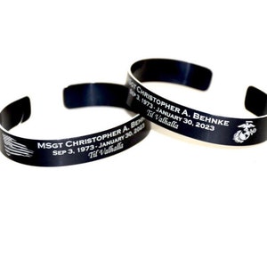 May include: Black metal bracelet with an engraved inscription that reads "MSGT CHRISTOPHER A. BEHNKE SEP 3, 1973-JANUARY 30, 2023 Til Valhalla" and a US Marine Corps emblem.